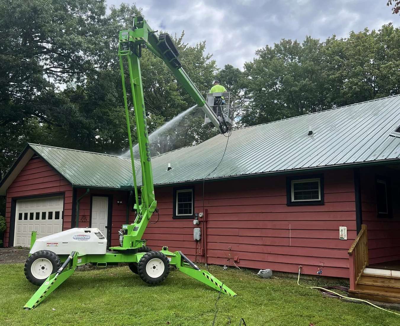 A tracked boom lift on a lawn with the basket raised over the metal roof of a red barn style building